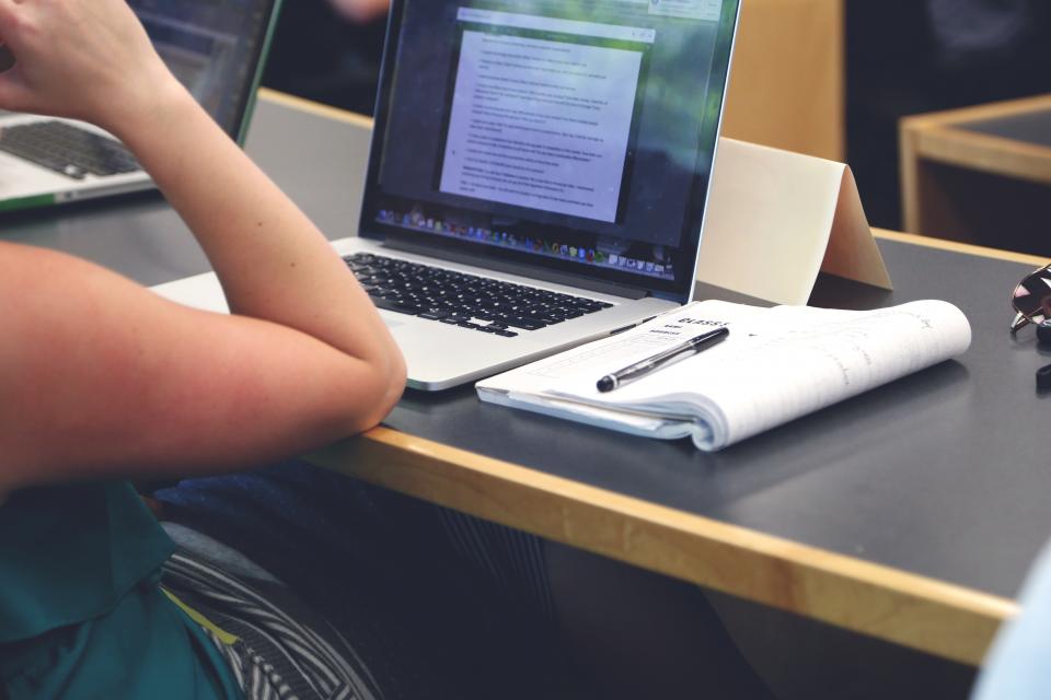 A student working through coursework on a laptop with a notebook open beside it